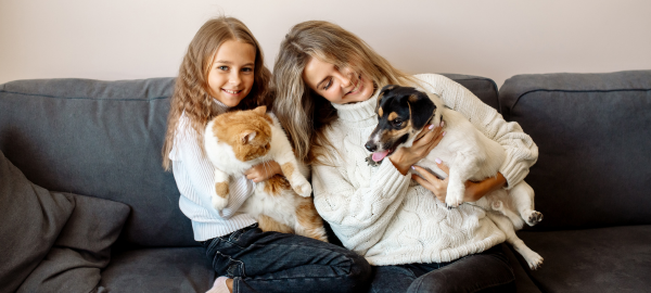 Ein Mädchen hat eine Katze auf dem Arm und eine Frau einen Hund und beide sitzen auf einem grauen Sofa